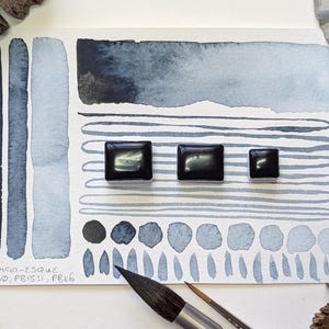May include: Watercolor paint swatches in shades of blue and gray on white paper. The image includes three black square paint pans, a paintbrush, and the text "INDIGO-ESQUE PB60, PB15:1, PBK6".