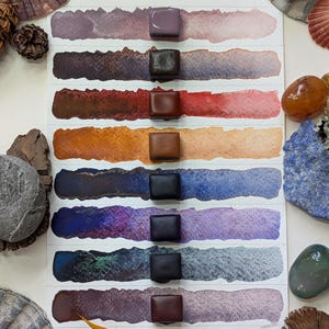 Könnte beinhalten: Ein Set aus acht rechteckigen Aquarellfarbnäpfchen, die in einer vertikalen Spalte auf einem weißen Papierbogen angeordnet sind, jeweils mit einem horizontalen Farbabstrich des entsprechenden Pigments von Mauve bis Dunkelbraun.