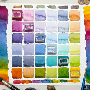 May include: A watercolor paint swatch chart with a grid of colorful squares and vertical color gradients. The squares display various hues, including pinks, blues, greens, and yellows. Paintbrushes and small rocks are visible.