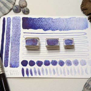 May include: Three watercolour paint pans in a light brown container with purple paint swatches on a white paper background. The paint pans are labelled "Stellar Glow", "Violet Glaze", and "Air Blue Gray".