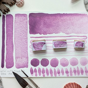 May include: A watercolor paint swatch card featuring various shades of red-violet. The card includes brush strokes, gradients, and circular and teardrop-shaped samples. Three small wrapped paint blocks are labeled "NEW ULTRAMARINE RED PR259" and "PR257".