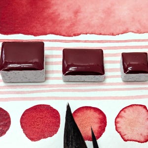 May include: Three square watercolor paint pans in varying sizes, filled with a deep burgundy color, sit on a white and pink striped surface. Below, are several swatches of the same color, along with a paintbrush.