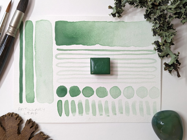 May include: Watercolor paint swatches in various shades of green, including a solid square of dark green paint. The word "FRITILLARY LEAF" is written on the paper. Paintbrushes and decorative elements are also visible.