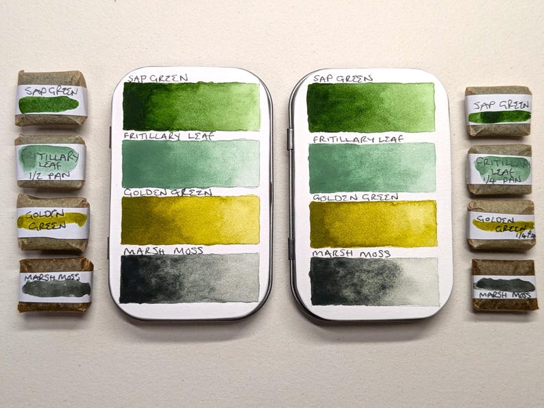 May include: Two silver watercolour palettes with green, yellow, and grey paint swatches labelled "Sap Green," "Fritillary Leaf," "Golden Green," and "Marsh Moss." Small wrapped watercolour pans are arranged around the palettes, also labelled.