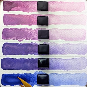 May include: A watercolor paint palette with six swatches of purple and blue hues. Each swatch is a gradient, transitioning from a darker shade to a lighter one. Square, dark paint blocks sit above each swatch. A paintbrush and other art supplies are visible.