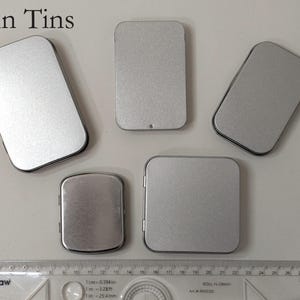May include: An assortment of five plain, silver-colored metal tins in various rectangular and square shapes. The text "Plain Tins" is at the top. A ruler is at the bottom, with measurements in inches and centimeters.