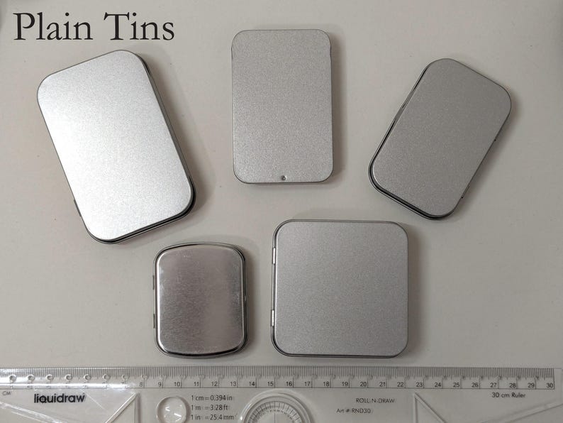 May include: An assortment of five plain, silver-colored metal tins in various rectangular and square shapes. The text "Plain Tins" is at the top. A ruler is at the bottom, with measurements in inches and centimeters.