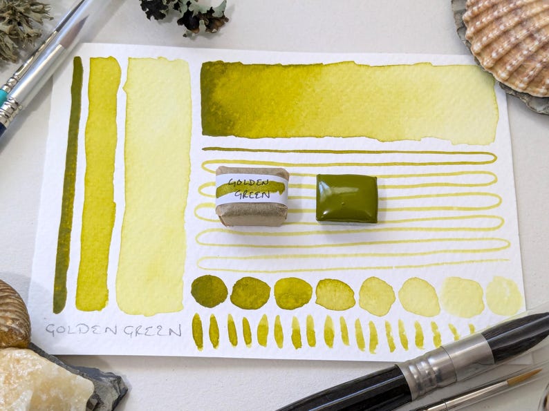 May include: Watercolor paint swatches in shades of golden green on white paper. The paper includes a watercolor paint block and a wrapped paint block labeled "GOLDEN GREEN". Paint swatches and a watercolor brush are also visible.