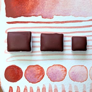 Könnte beinhalten: Drei quadratische Aquarellfarben in verschiedenen Größen, gefüllt mit einem satten, dunkelbraunen Pigment. Die Farbtöpfe liegen auf Aquarellpapier mit roten und rosa Aquarellfarben und Streifen.