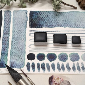 Peut inclure: Un coffret de peinture aquarelle avec trois godets de peinture noire. La peinture est disposée en rangée sur une surface blanche. La peinture est entourée d'échantillons de peinture dans différentes nuances de bleu et de gris. Le texte "Lights in New Batch" est écrit sur la surface.