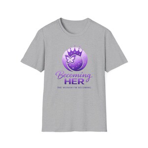May include: A gray t-shirt featuring a purple graphic with a crown, butterfly, and the words "Becoming Her" and "The Woman I'm Becoming". The design is centered on the front of the short-sleeved shirt.
