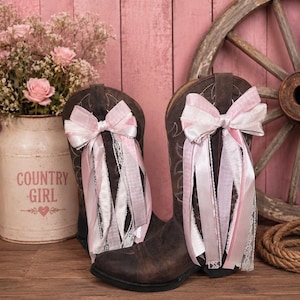 Op de afbeelding: Een paar donkerbruine leren cowboylaarzen met decoratieve strikken van gelaagde roze, witte en geruite linten met kanten randen aan de schachten. De laarzen zijn voorzien van klassieke westernstiksels op het donkere leer.