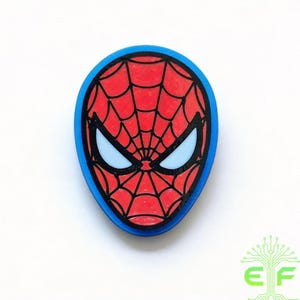 May include: A Spiderman-themed emblem featuring a red mask with a black web design, blue eyes, and a blue border. The design is a close-up of the superhero's face, with a white background.