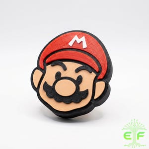 May include: A 3D-printed figure of Mario's head, featuring a red cap with a white "M", a tan face, black mustache, and black outline. The figure is a recognizable character from the Super Mario video game series.