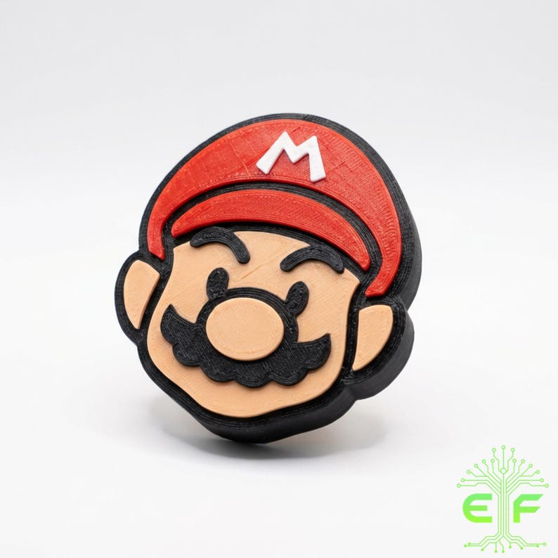 May include: A 3D-printed figure of Mario's head, featuring a red cap with a white "M", a tan face, black mustache, and black outline. The figure is a recognizable character from the Super Mario video game series.