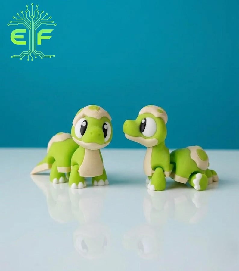 May include: Two green and tan cartoon turtle figurines with large black eyes and white highlights. The turtles are on a reflective surface against a teal background. The logo "EF" is in the top left corner.