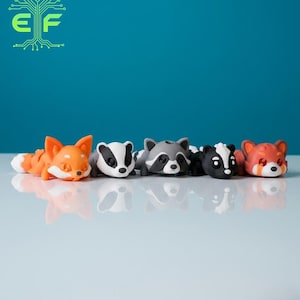 May include: A collection of five animal figurines: a fox, a badger, a raccoon, a skunk, and a red panda. Each figurine is lying down, with detailed features and vibrant colors. The background is a solid teal color, and the figurines are on a white surface.