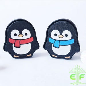 May include: Two penguin-shaped decorative items. Each penguin has a black body, white belly, and a colorful scarf. One penguin has a red scarf, and the other has a blue scarf. The items are on a white background.