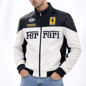 May include: A black, white, and yellow Ferrari racing jacket with the Ferrari logo and text. The jacket has a zip-up front, black cuffs, and a stand-up collar. The jacket is worn with blue jeans.