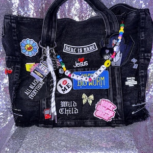 May include: A black denim tote bag adorned with various patches and charms. Patches include text like Real Is Rare, I Love Jesus, Big Worm, Wild Child, and Make Today Great. A beaded chain with a small duck and star beads hangs from the handle.