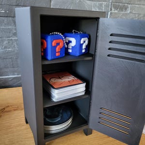 May include: A small, dark grey locker with an open door, revealing two shelves. The top shelf holds two blue cube-shaped keychains with question marks. The middle shelf has a stack of cards. The bottom shelf has a stack of plates.