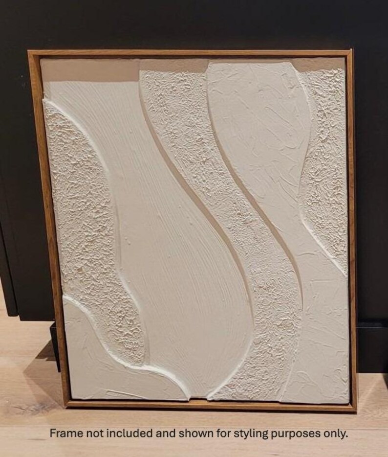 May include: Abstract white textured art piece with a wooden frame. The artwork features flowing, curved shapes with varying textures. The frame is a warm, natural wood tone. The text at the bottom reads: Frame not included and shown for styling purposes only.