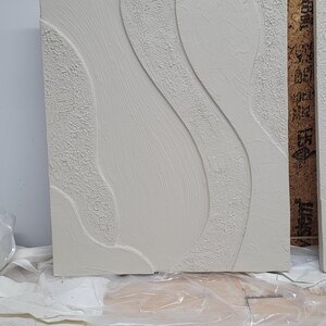 May include: A rectangular, off-white textured wall art piece with a flowing, abstract design. The artwork features raised, curved lines and textured areas, creating a tactile and visual contrast. The piece is likely for home decor.