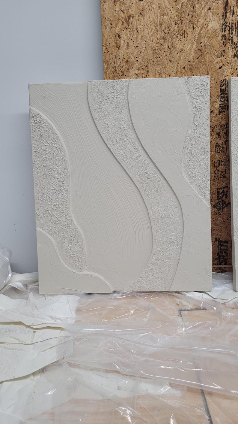 May include: A rectangular, off-white textured wall art piece with a flowing, abstract design. The artwork features raised, curved lines and textured areas, creating a tactile and visual contrast. The piece is likely for home decor.