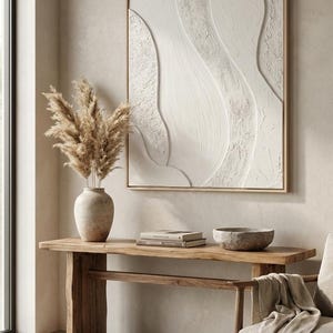 May include: A neutral-toned interior scene featuring a large, textured white abstract art piece in a gold frame. A wooden console table holds a vase with dried pampas grass, books, and a stone bowl. A beige throw blanket drapes over a chair.