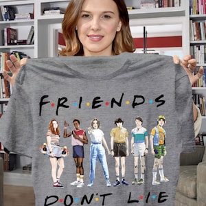 May include: Gray t-shirt featuring the text "FRIENDS DON'T LIE" in black, with colorful dots above the word "FRIENDS". The shirt displays a graphic of characters from a popular TV show. The characters are in various outfits.
