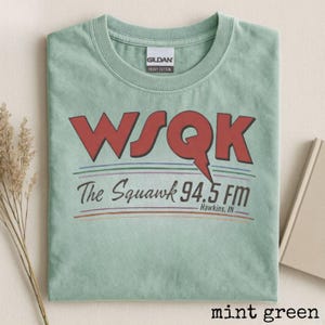 Puede incluir: Camiseta verde menta con el texto "WSQK The Squawk 94.5 FM Hawkins, IN" en letras rojas y negras. La camiseta es de algodón grueso y tiene cuello redondo.