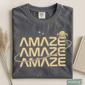 Könnte beinhalten: Ein dunkelgraues T-Shirt mit dem Wort "AMAZE", das dreimal in einer gelben Retro-Schrift gestapelt ist. Eine gelbe Grafik einer Weltraumkreatur und eine gebogene Linie befinden sich über und unter dem Wort. Das Shirt hat ein Comfort Colors-Etikett.