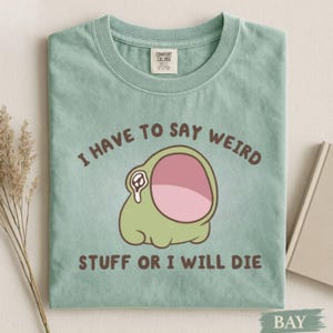 Puede incluir: Camiseta verde claro con el texto "I HAVE TO SAY WEIRD STUFF OR I WILL DIE" en marrón. La camiseta presenta una rana de dibujos animados con una gran boca abierta y una lágrima. Marca Comfort Colors.
