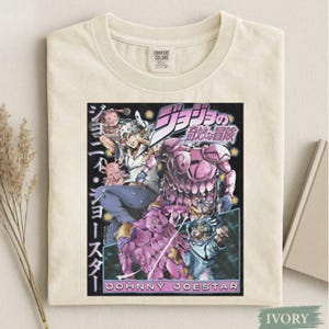 Puede incluir: Camiseta de color marfil con un gráfico colorido de la serie de anime "JoJo's Bizarre Adventure". El diseño incluye personajes y texto japonés, con el nombre "JOHNNY JOESTAR" en la parte inferior. La camiseta tiene una etiqueta Comfort Colors.