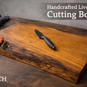 Könnte beinhalten: Handgefertigtes Schneidebrett mit natürlicher Kante. Ein schwarzes Messer liegt auf dem Brett, zusammen mit einer Tomate und Kräutern. Das Brett ist etwa 46 cm lang und 30 cm breit.