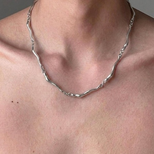 Könnte beinhalten: Eine silberfarbene Halskette bestehend aus einer Reihe miteinander verbundener, glatter, wellenförmiger Metallglieder. Die Kette hat eine polierte, reflektierende Oberfläche und zeigt ein fließendes, organisches Design mit gleichmäßigen, geschwungenen Segmenten.