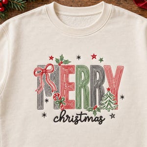 May include: A cream-colored crewneck sweatshirt featuring embroidered text that reads MERRY in large, multi-colored block letters with a red bow, a candy cane, a green Christmas tree, and holly leaves, with the word christmas in black script.