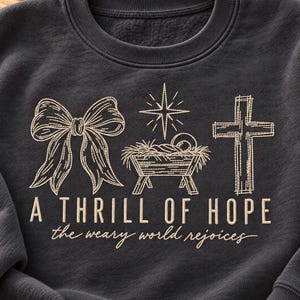 May include: A dark charcoal gray crewneck sweatshirt featuring a light beige embroidered design of a bow, a manger scene with a star, and a cross. Below the graphics, the text reads A THRILL OF HOPE and the weary world rejoices.