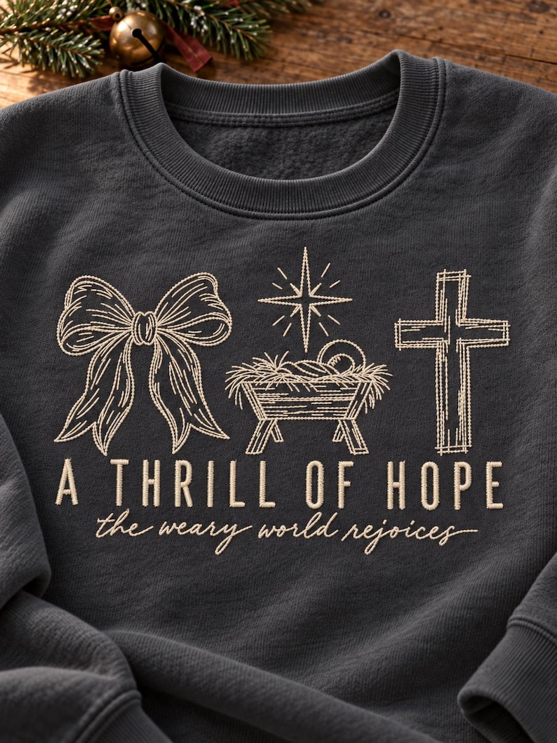 May include: A dark charcoal gray crewneck sweatshirt featuring a light beige embroidered design of a bow, a manger scene with a star, and a cross. Below the graphics, the text reads A THRILL OF HOPE and the weary world rejoices.