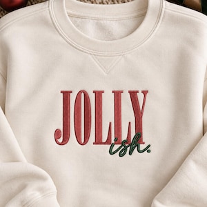 May include: A cream-colored crewneck sweatshirt featuring embroidered text on the chest. The word JOLLY is stitched in red capital letters, with the word ish. embroidered in dark green lowercase script letters positioned below the right side.