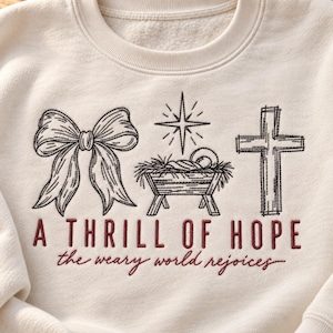 May include: An off-white crewneck sweatshirt featuring black embroidered line art of a bow, a manger with a star above it, and a cross. Below the graphics, the text reads A THRILL OF HOPE in block letters and the weary world rejoices in script.