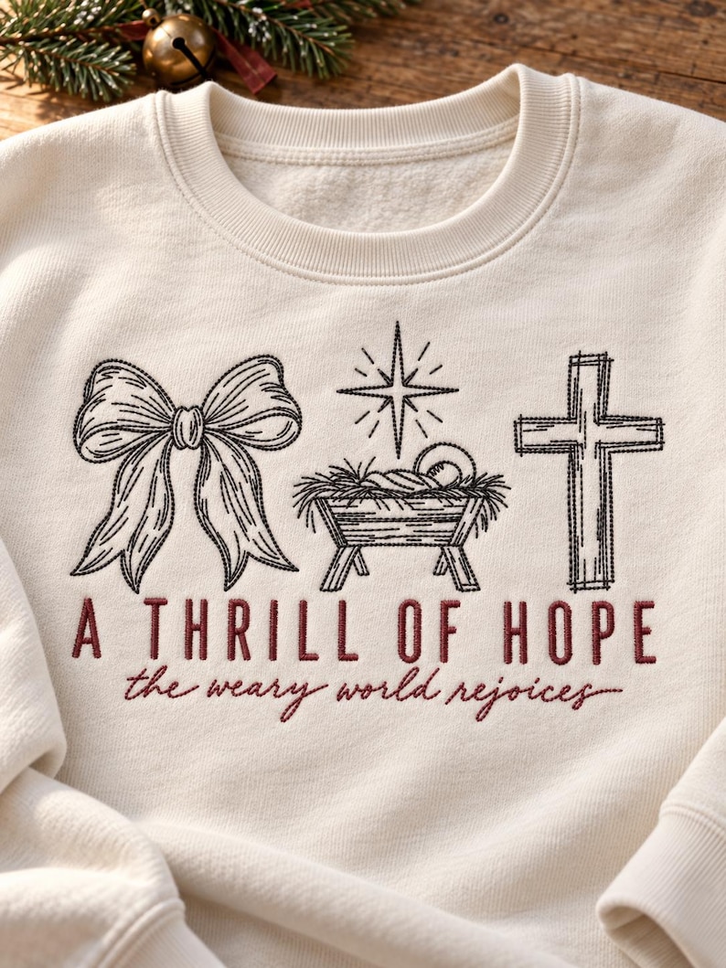 May include: An off-white crewneck sweatshirt featuring black embroidered line art of a bow, a manger with a star above it, and a cross. Below the graphics, the text reads A THRILL OF HOPE in block letters and the weary world rejoices in script.