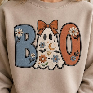 Peut inclure: Un sweat-shirt beige à col rond avec une broderie formant le mot BOO. La lettre B est bleue avec des motifs floraux, la lettre O est orange avec des fleurs, et le fantôme central porte un nœud orange avec des motifs de fleurs, de chauves-souris et de lune.