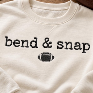 Può includere: Una felpa girocollo color crema con testo ricamato in nero che recita bend & snap, posizionato sopra una piccola icona di un pallone da football americano ricamata in nero. Il capo presenta un colletto a coste con cucitura triangolare.