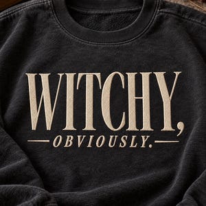 May include: A black crewneck sweatshirt featuring the embroidered text WITCHY, OBVIOUSLY. in a light beige thread. The garment has a washed, vintage-style finish with a ribbed collar and cuffs, showcasing a clean, centered design on the chest.