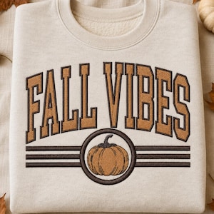 May include: A beige crewneck sweatshirt featuring the embroidered text FALL VIBES in a brown collegiate font. Below the text, a circular emblem contains an orange pumpkin graphic, flanked by three horizontal brown lines on each side.