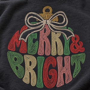 May include: A charcoal gray sweatshirt featuring an embroidered Christmas ornament design. The ornament is shaped like a circle with a bow on top and contains the text MERRY & BRIGHT in red, green, and gold thread with a spiral stitch pattern.