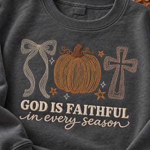 Pode incluir: Uma camisola de gola redonda cinzento-carvão com um design bordado de um laço, uma abóbora e uma cruz. O texto "GOD IS FAITHFUL in every season" está cosido abaixo dos gráficos numa combinação de letras de forma e cursivas.