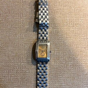 May include: A silver-toned wristwatch with a rectangular gold-colored face and a silver link band. The watch face displays the number 12 and two smaller dials. The watch band is made of small, rectangular links.