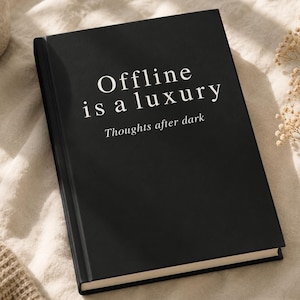 Op de afbeelding: Een zwart notitieboek met harde kaft en witte tekst op de voorkant die luidt "Offline is a luxury", gevolgd door de kleinere tekst "Thoughts after dark". Het boek heeft een strakke, rechthoekige vorm met een matte afwerking en witte pagina's.